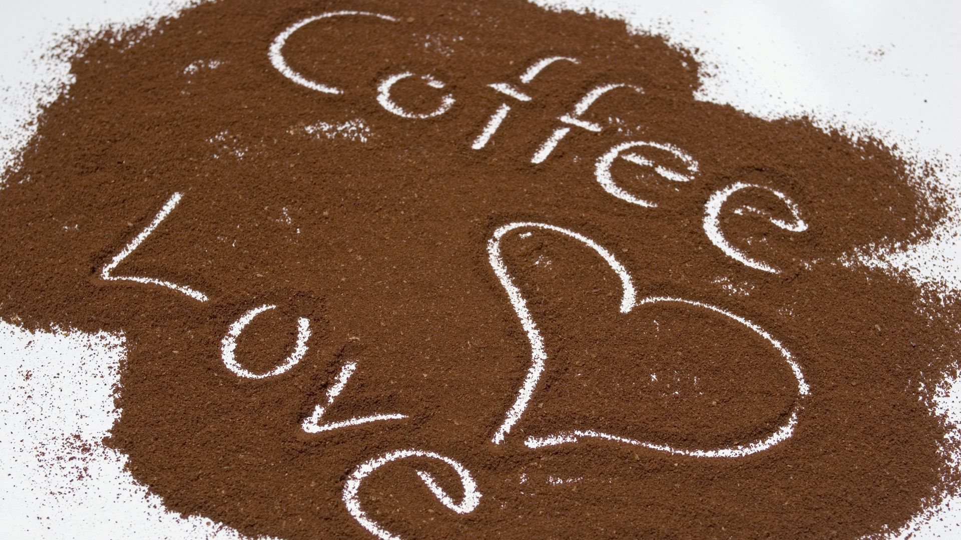 A white surface covered in a pile of brown coffee grounds. The words "Coffee Love" are written in the grounds, with the word "Love" containing a heart shape.
