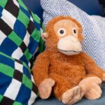 A plush, brown stuffed monkey sits nestled between two pillows on a blue surface. The pillow on the left has a green, black, and blue diamond/harlequin pattern, and the pillow behind the monkey has a white and blue chevron/herringbone pattern.