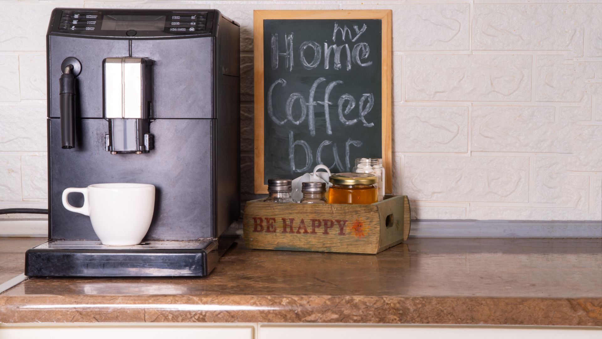 A home coffee bar setup with a black espresso machine, a white coffee cup, and a small crate of honey and other ingredients. A small chalkboard sign reads "My Home Coffee Bar."