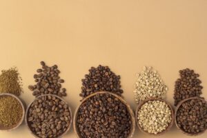An overhead shot of different types of coffee beans and grounds arranged in wooden bowls on a light brown surface. The types shown include light, medium, and dark roasted beans, green beans, and ground coffee.