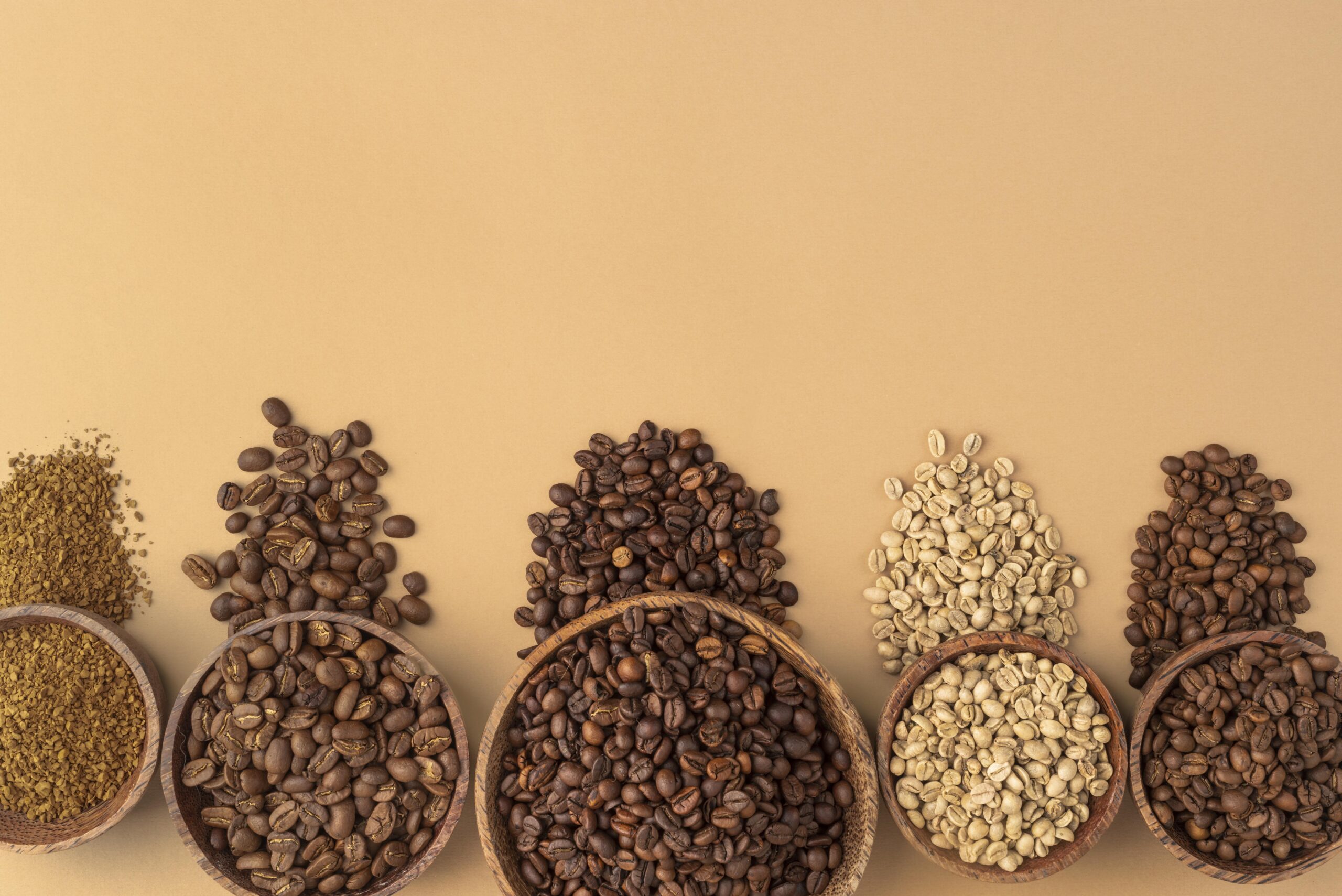 An overhead shot of different types of coffee beans and grounds arranged in wooden bowls on a light brown surface. The types shown include light, medium, and dark roasted beans, green beans, and ground coffee.