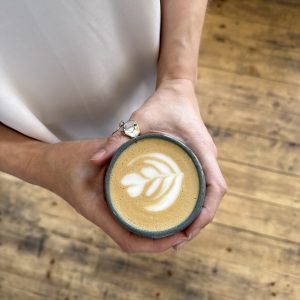A top-down view of two hands holding a ceramic mug of latte with simple, white tulip latte art. The person is wearing a light-coloured garment, and the background is a rustic wooden floor.