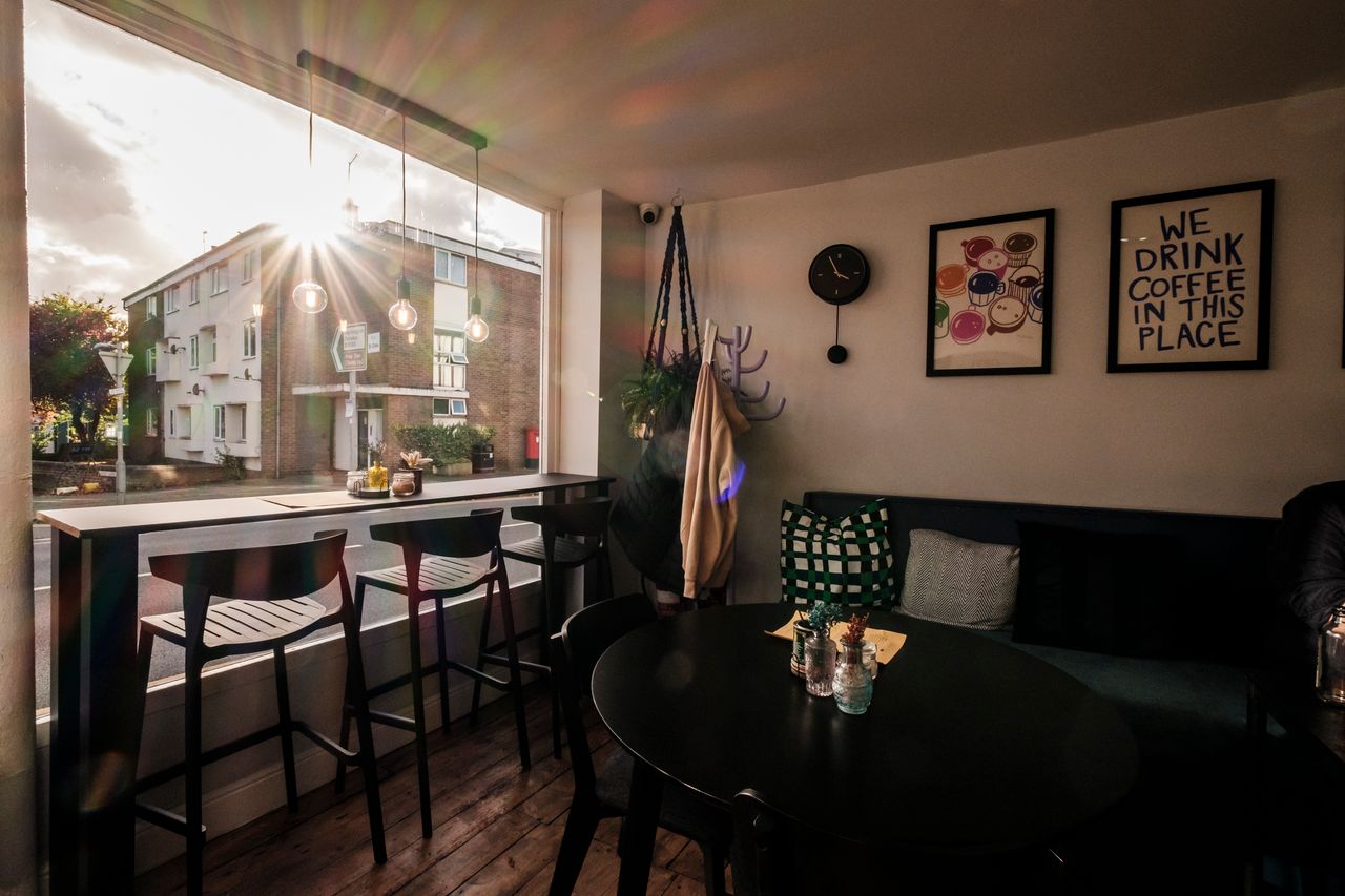Interior shot of a café with sunlight streaming through a large window overlooking the street, showing the bar seating and a round table.