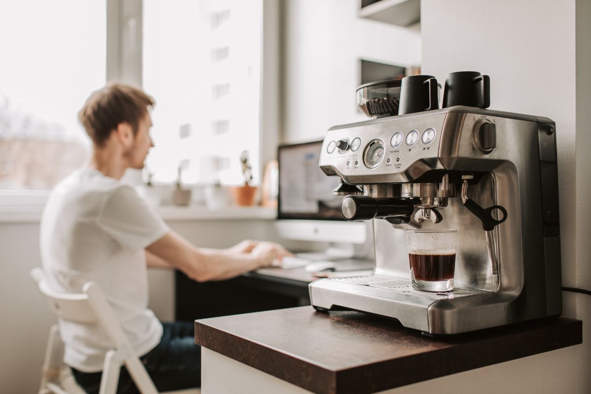 A stainless steel espresso machine sits on a kitchen counter in the foreground, while a person in a white t-shirt sits at a desk working on a computer in the blurred background.