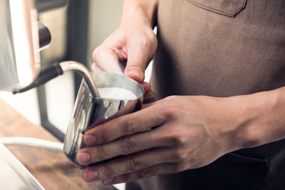 A close-up shot of a person wearing a brown apron using a steam wand to froth milk in a stainless steel pitcher.
