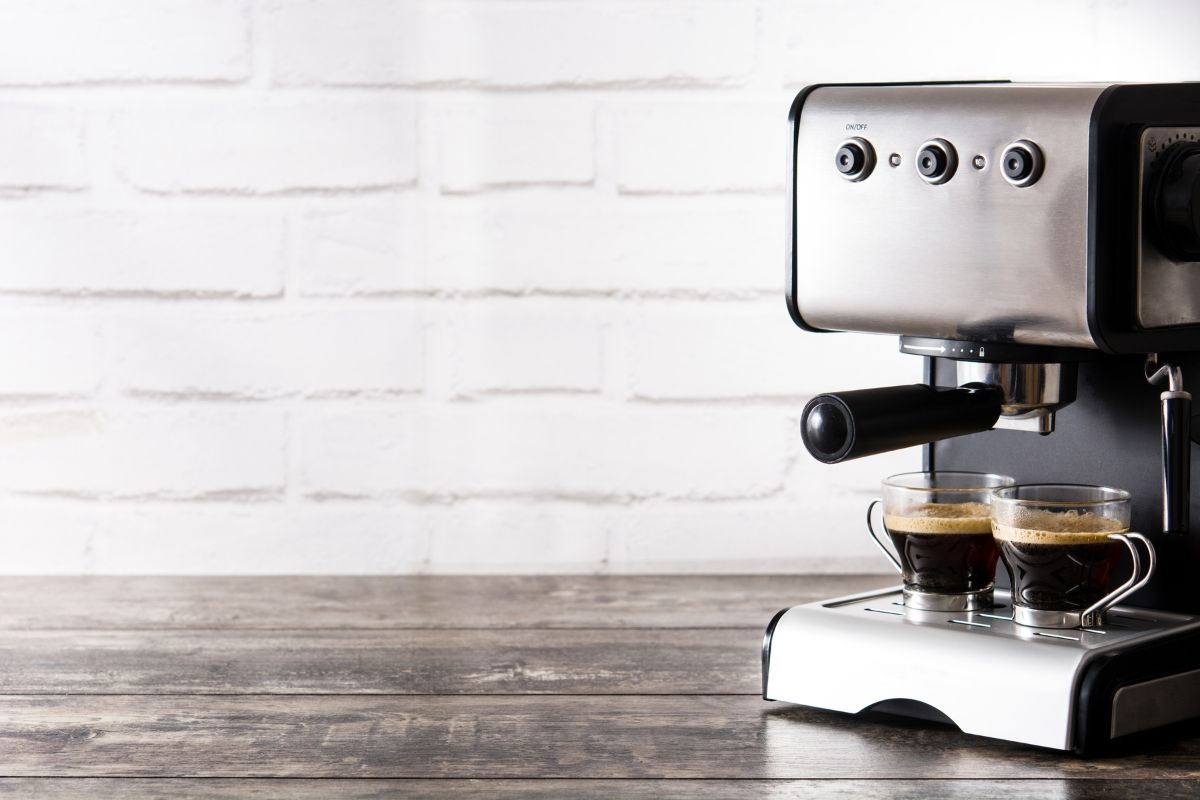 A close-up of a silver espresso machine brewing two small glass cups of coffee, set against a white brick wall and a dark wooden surface.