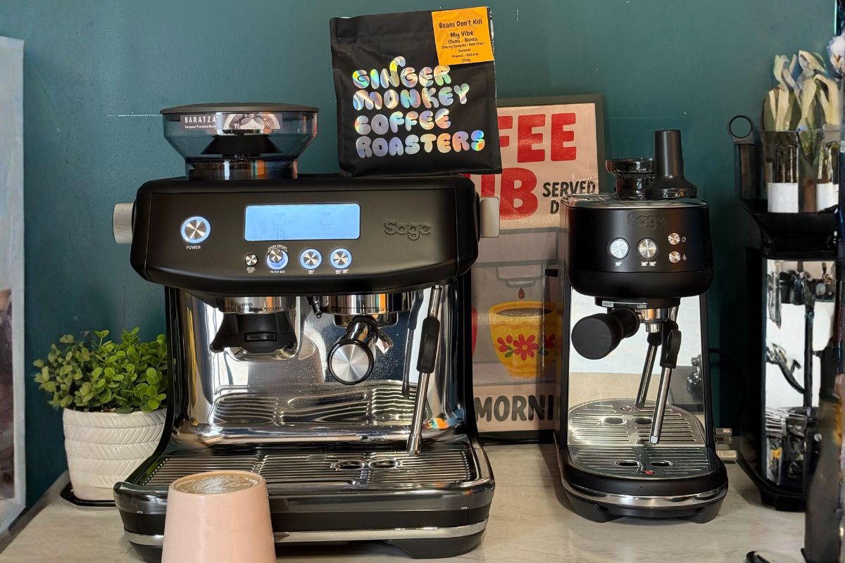 Two different models of black Sage espresso machines stand side-by-side on a counter, with a bag of coffee beans resting on top of the larger machine.