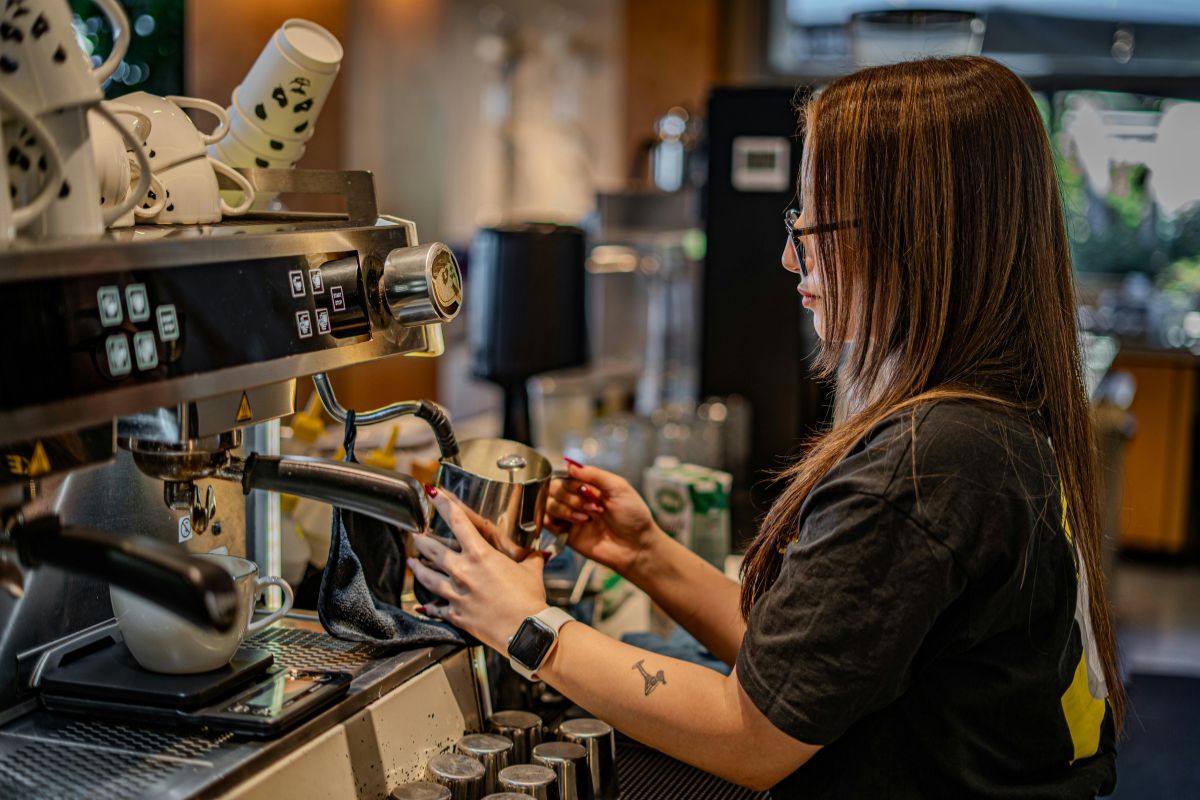 A person with long hair and glasses stands at a large professional espresso machine, holding a metal pitcher to steam milk in a cafe setting.