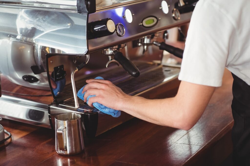 A person in a white t-shirt uses a blue cloth to wipe down the drip tray area of a stainless steel espresso machine.