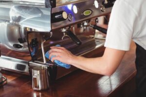 A person in a white t-shirt uses a blue cloth to wipe down the drip tray area of a stainless steel espresso machine.