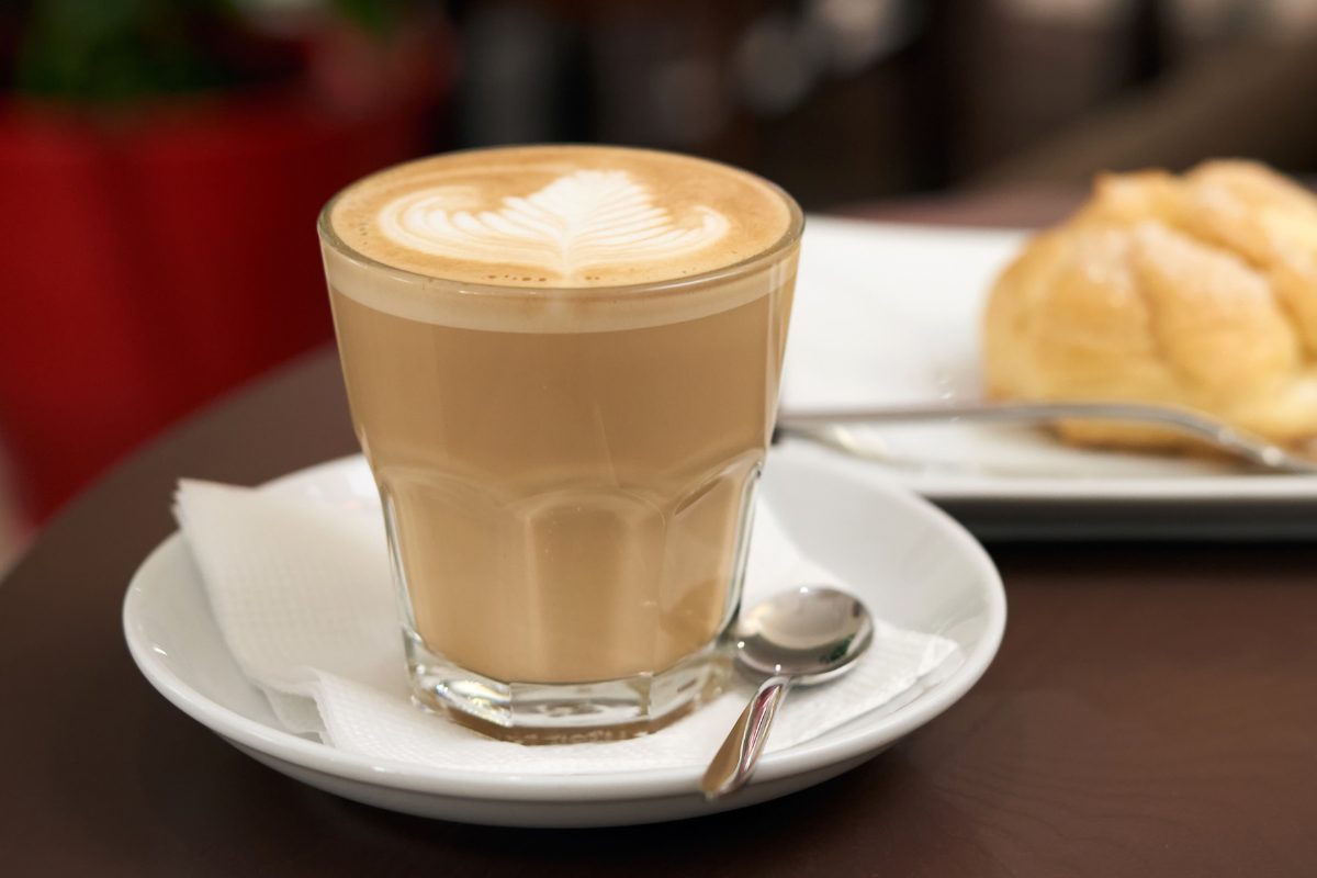 A flat white coffee in a clear glass with detailed rosette latte art, sitting on a white saucer with a spoon.