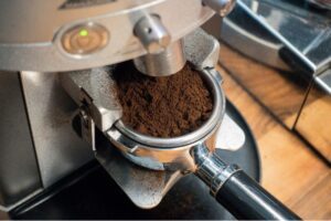 Freshly ground coffee filling an espresso portafilter directly under a commercial burr grinder chute.